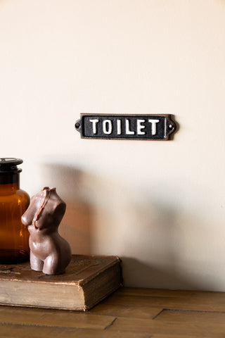 The Black & White Toilet Door Hanging Sign displayed on a neutral wall, styled above a wooden surface with a book, glass jar and candle.