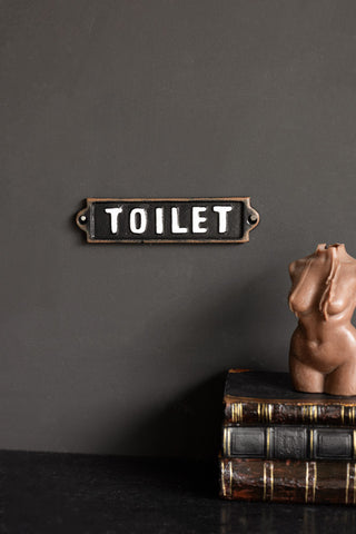 The Black & White Toilet Door Hanging Sign displayed on a black wall, above a black surface styled with books and a candle in the shape of a female figure.