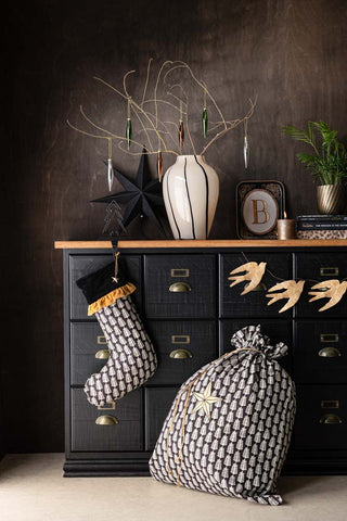 The Block Print Christmas Stocking hanging from a black and wooden storage unit, styled with a Christmas sack, garland and various other decorative accessories.