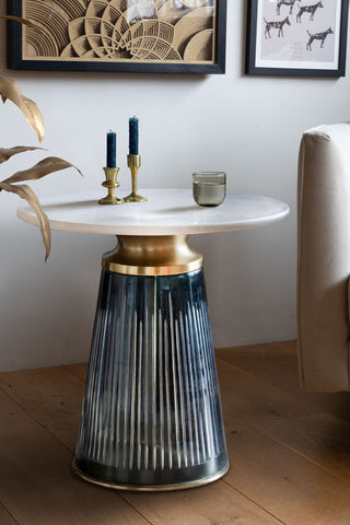 The Blue Tapered Glass & Marble Side Table styled next to a sofa and large plant, with some candlestick holders and a glass of water on.