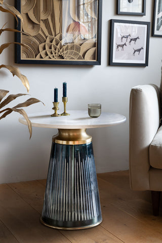 The Blue Tapered Glass & Marble Side Table next to a sofa and large plant, with a glass of water and some candlestick holders on.