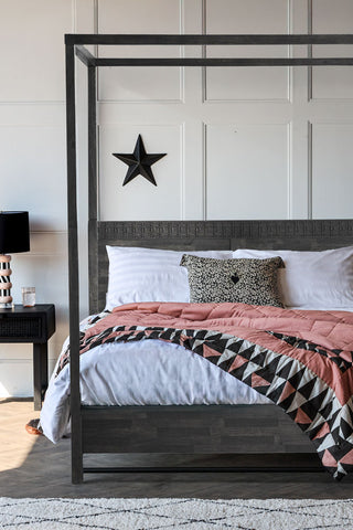 The Boho-Inspired Boutique Four Poster Bed styled in a bedroom with a cushion and pink throw on, with a rug, bedside table, lamp, glass of water and star decoration on the wall.