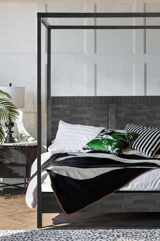 The Boho-Inspired Boutique Four Poster Bed styled with various cushions and a throw on, with a bedside table, lamp and plant to the side of the shot.