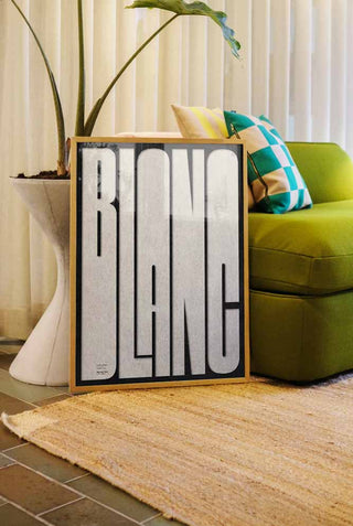 The Bold Blanc Art Print in a frame styled in a living room, leaning against a sofa and a plant.