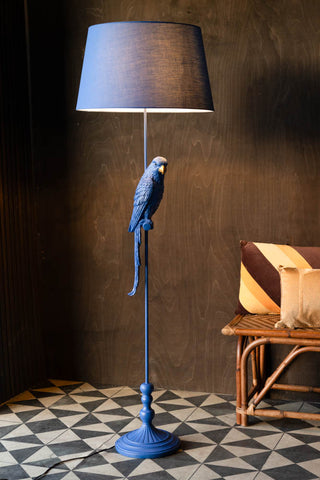 The Bold Blue Parrot Floor Lamp with Shade switched on and styled in a room with a bench with some cushions.