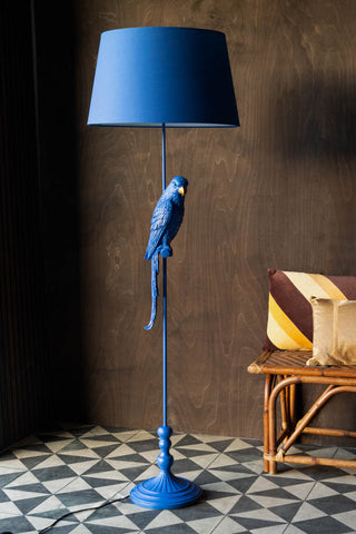 The Bold Blue Parrot Floor Lamp with Shade styled with a bench and some cushions, on a geometric patterned floor in front of a wooden wall.