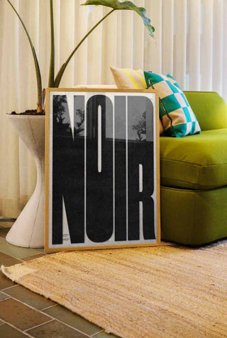 The Bold Noir Art Print styled in a frame in a living room, leaning against a sofa and a plant.