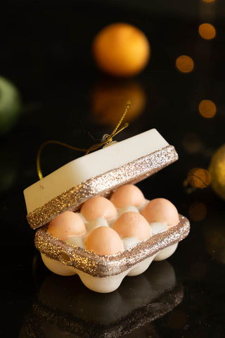 The Box Of Eggs Christmas Tree Decoration displayed on a dark surface with baubles and fairy lights in the background.