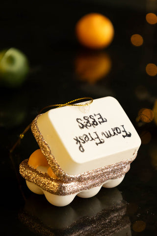 The Box Of Eggs Christmas Tree Decoration seen from the reverse side, styled on a dark surface with baubles and fairy lights in the background.