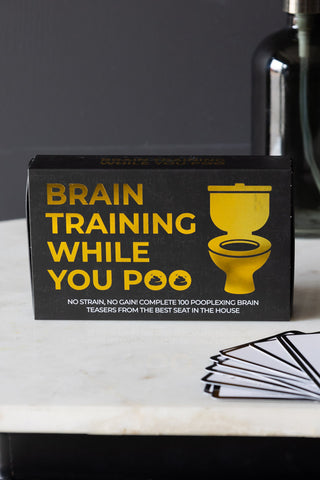 The box of the Brain Training While You Poo Card Pack displayed with some cards and a soap dispenser.