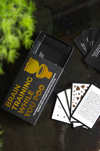 The Brain Training While You Poo Card Pack with some of the cards loose, displayed on a dark surface with some greenery.