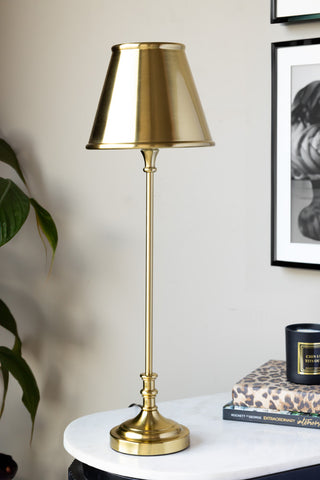 The Brass Slim Table Lamp styled on a marble table with some books and a candle, with a plant and art prints in the background.