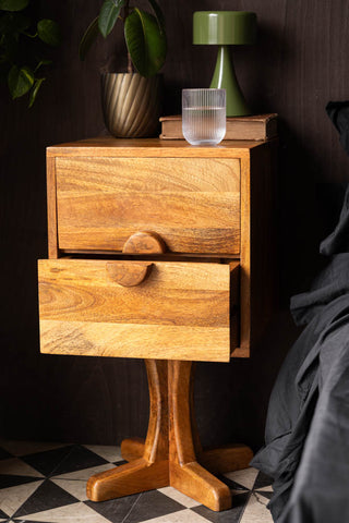 The Brown Wooden Footed Bedside Table with the bottom drawer open, with a glass, book, plant and lamp on, styled next to a bed.