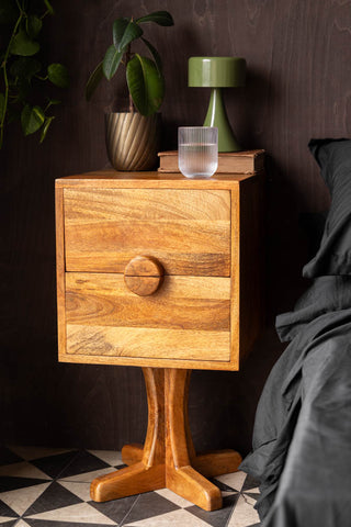 The Brown Wooden Footed Bedside Table styled next to a bed, with a drinking glass, plant, book and lamp on.