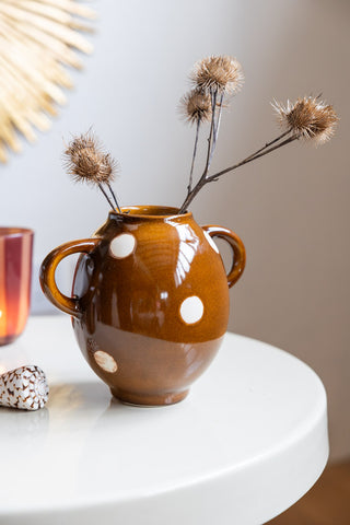 The Brown & Ivory Dotted Display Vase styled with some flowers inside, on a round white table with some other accessories.