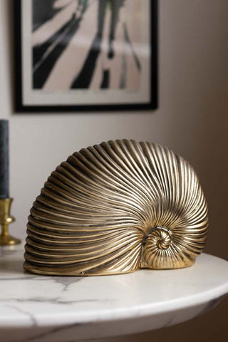 Lifestyle image of the Brushed Gold Faux Sea Shell Ornament on a white marble table, with a candlestick holder and art print in the background.