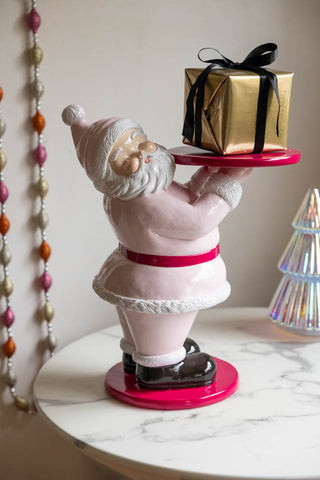 The Butler Santa In Pink Christmas Decoration styled on a white marble table with a present on, next to a Christmas tree ornament and a Christmas garland.