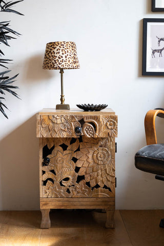 The Carved Floral Design Wood Bedside Table styled with a lamp and trinket tray with the drawer open.