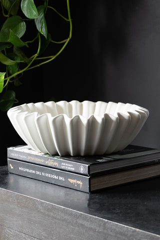 The White Ceramic Waved Bowl styled on a stack of books on a black sideboard, with a plant in the background.