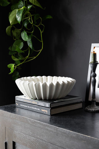 A white wavy bowl displayed on a stack of books, on a black sideboard with a lit candle, art print and some greenery in the background.