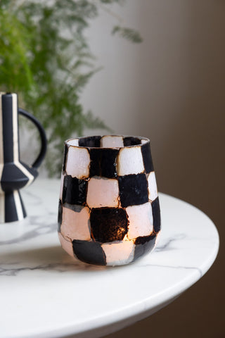 The Checkered Black & White Glass Tealight Holder with a lit tealight inside, displayed on a round marble table with a vase and plant in the background.