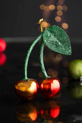 The Cherries Christmas Tree Decoration styled on a black surface with baubles and fairy lights in the background.