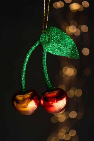 The Cherries Christmas Tree Decoration hanging in front of a dark wall with fairy lights in the background.