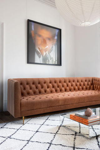 The Chesterfield Antique Brown Leather 3-Seater Sofa styled with a rug, coffee table with accessories on and a large framed artwork on the wall.