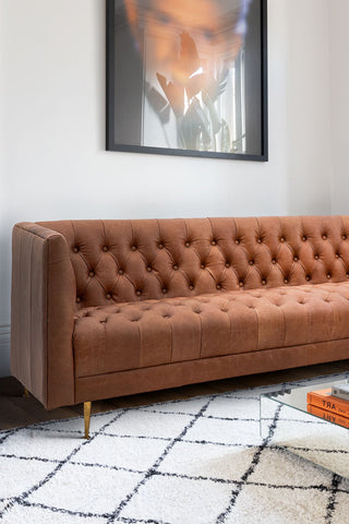 The Chesterfield Antique Brown Leather 3-Seater Sofa styled on a rug behind a coffee table, with a large art print on the wall above.