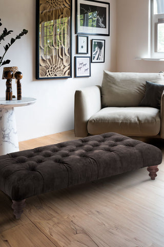 The Chesterfield Charcoal Buttoned Footstool styled in a living room in front of and armchair and table.