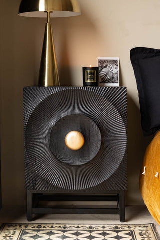 The Circular Ribbed Wood Detail Bedside Table styled with a lamp, candle and photograph on, next to a bed.