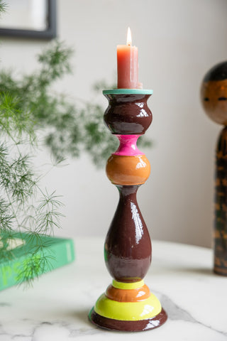 The Colourful Candlestick Holder in Chocolate & Orange with a lit candle inside, with a plant, book and ornament in the background.