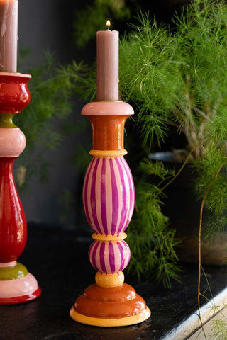 The Colourful Candlestick Holder in Pink Stripes styled with a lit candle inside, with another candlestick holder and a plant in the background.