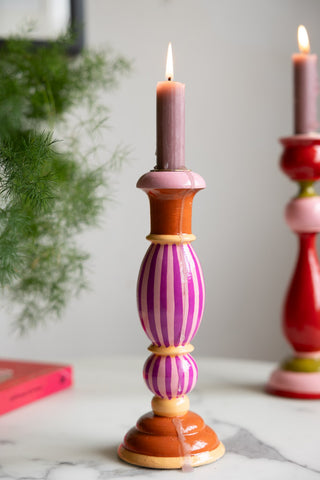 The Colourful Candlestick Holder in Pink Stripes with a lit candle inside, styled with another colourful candlestick holder, a book and a plant.