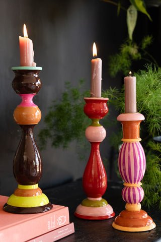 The Colourful Candlestick Holder in Red & Pink styled with a lit candle inside alongside two other colourways, some books and a plant.