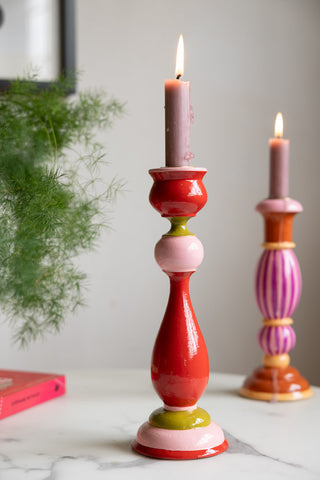 The Colourful Candlestick Holder in Red & Pink styled with a lit candle inside, with another candlestick holder, book and plant in the background.