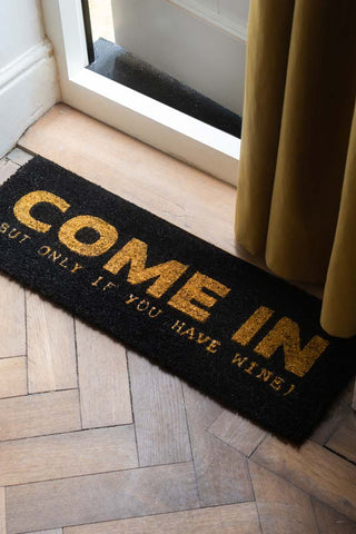 The Come In (But Only If You Have Wine) Coir Doormat styled in a doorway with a door curtain.