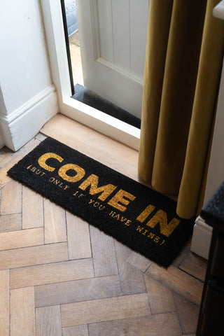 The Come In (But Only If You Have Wine) Coir Doormat displayed on a doorway, with a door curtain hanging above it.