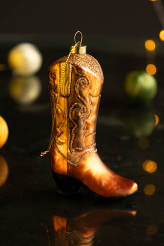 The Cowboy Boot Christmas Tree Decoration displayed on a dark surface with baubles and glowing lights in the background.