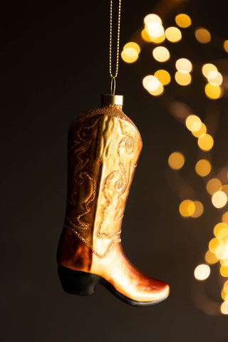 The Cowboy Boot Christmas Tree Decoration hanging in front of a dark wall wit lights glowing in the background.