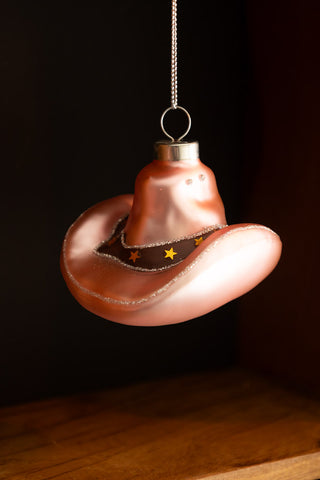 The Cowgirl Hat Christmas Tree Decoration hanging in front of a wooden surface.