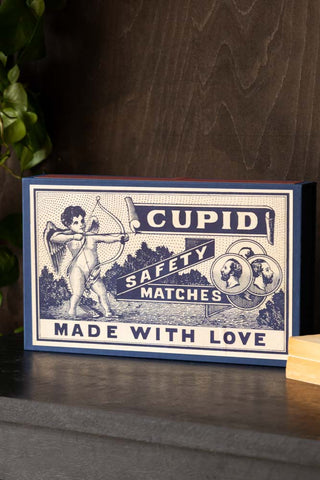 The Cupid, Made With Love Giant Luxury Matchbox standing on a dark sideboard, styled with some books and a plant.