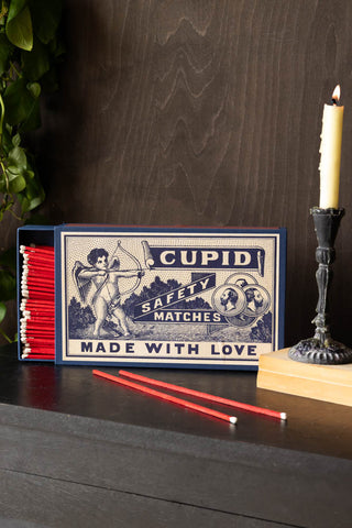 The Cupid, Made With Love Giant Luxury Matchbox open with some matches loose, styled with some books, a lit candle and a plant.