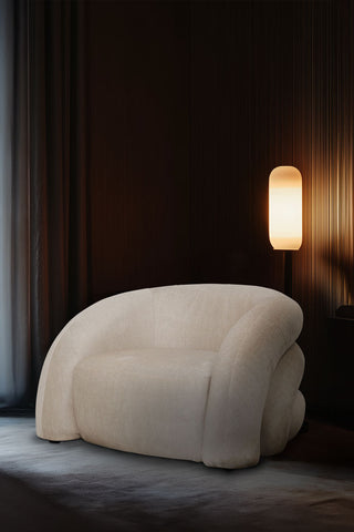 The Curved Cream Snug Armchair styled in the corner of a dark room with a curtain and a lamp.