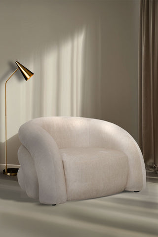 The Curved Cream Snug Armchair styled in front of a neutral wall with a gold lamp.