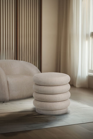 The Curved Cream Snug Footstool styled on a rug in front of an armchair in a neutral living room.