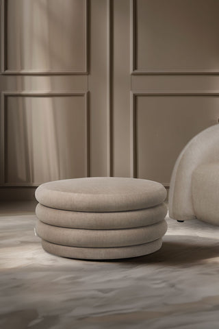 The Curved Cream Snug Ottoman styled in a living room with an armchair and a panelled neutral wall in the background.