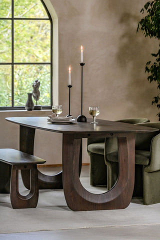 The Curved Mango Wood Dining Table styled with tableware and lit candles on, styled with a bench, dining chairs, ornaments and a large plant in the background.