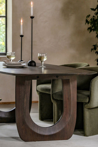 Close-up of the Curved Mango Wood Dining Table styled with glasses, plates and lit candles on, with some dining chairs and a large plant in the background.