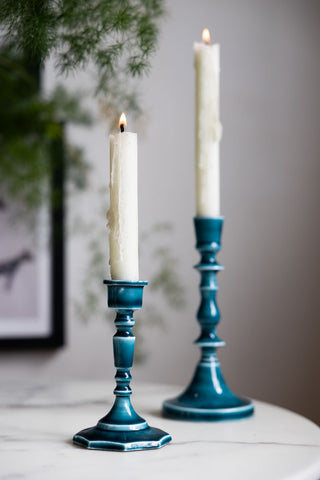 The Short and Tall Deep Blue Enamel Cast Style Candlestick Holders displayed with lit candles inside, styled on a white marble table with a plant and black frame in the background.